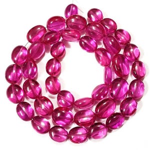 May include: A strand of oval-shaped, translucent, fuchsia-colored beads. The beads are strung together, creating a necklace-like arrangement. The beads have a glossy finish and are evenly sized, with a slight variation in shape.