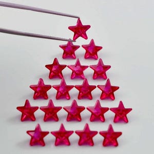 May include: A collection of vibrant, star-shaped gemstones arranged in a triangular pattern. The gems are a deep pink color and appear to be faceted, catching the light. A pair of tweezers holds one of the stars above the arrangement.