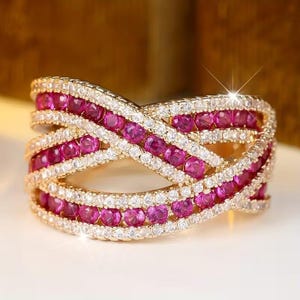 May include: A gold-toned ring with an interwoven design. The ring features rows of small, round, pink gemstones and clear, sparkling stones. The gemstones are set in a crisscross pattern, creating a visually appealing and intricate design.