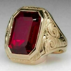 May include: A gold-colored ring featuring a large, rectangular, deep red gemstone. The ring has an ornate, engraved design around the gemstone and on the band, showcasing a classic, vintage style.