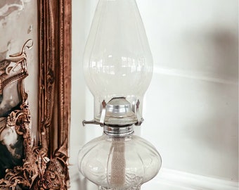 May include: A clear glass oil lamp with a decorative, textured base and a clear glass chimney. The lamp is sitting on a wooden surface.