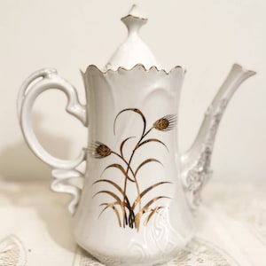 May include: White porcelain teapot with a gold lid and gold floral design. The teapot has a scalloped rim and a delicate handle.