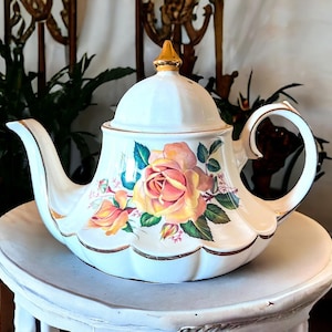 May include: A white ceramic teapot with a floral rose design. The teapot features a gold-colored handle, spout, and trim. The lid is topped with a gold finial. The teapot is sitting on a white, scalloped-edge table.