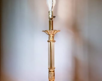 May include: A tall, ornate gold floor lamp with a white bulb. The lamp features a detailed base, a fluted column, and decorative accents. The lamp's design is classic and elegant, suitable for interior lighting.