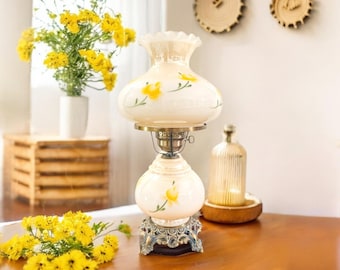 May include: A vintage-style cream-colored lamp with yellow floral accents. The lamp has a glass globe and a decorative base. The lamp is on a wooden table, with yellow flowers and a vase in the background. The lamp is a decorative home decor item.