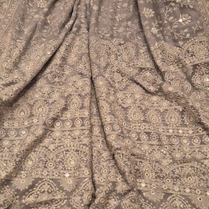 May include: A gray silk saree with intricate white embroidery and silver sequins. The saree has a border with a repeating pattern of flowers and leaves.