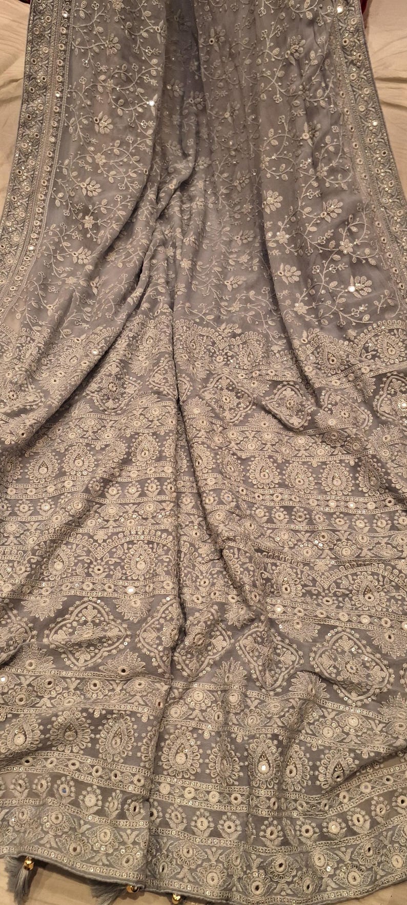 May include: A gray silk saree with intricate white embroidery and silver sequins. The saree has a border with a repeating pattern of flowers and leaves.