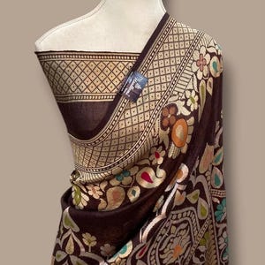 Puede incluir: Un textil marrón oscuro con intrincados patrones florales y geométricos. La tela está drapeada sobre un maniquí, mostrando el diseño detallado con acentos dorados, rojos y verdes. Una etiqueta es visible.