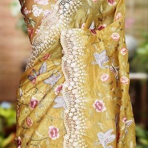 Puede incluir: Sari amarillo mostaza con bordados florales y de pájaros. El sari presenta un delicado borde de encaje blanco y está drapeado sobre un maniquí. El diseño incluye flores rosas y pájaros grises.