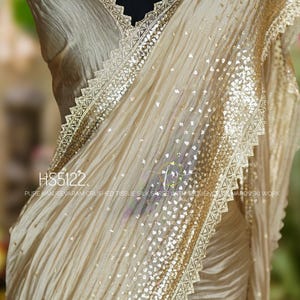 May include: A beige saree with gold sequin and lace trim. The fabric is draped over a mannequin, showcasing the texture and design. A three-strand necklace with large, clear stones is visible.