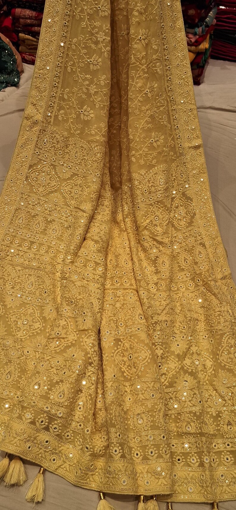 May include: A yellow chiffon saree with intricate embroidery and mirror work. The saree has a delicate floral pattern and a border with a geometric design. The saree is adorned with small, round mirrors that add a touch of sparkle.