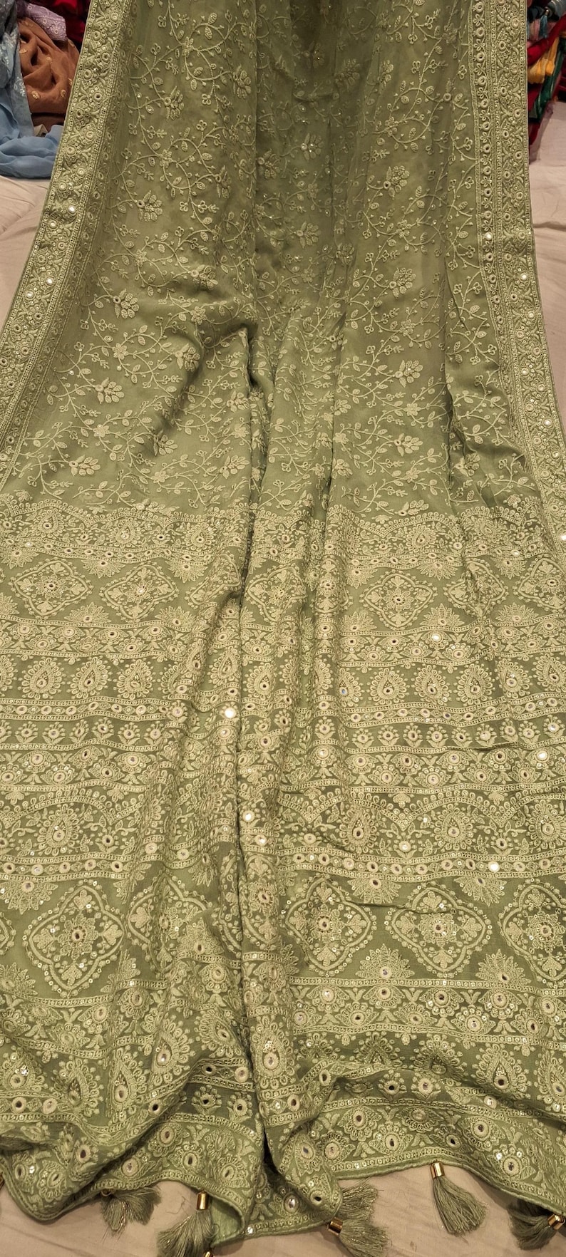May include: A sage green chiffon saree with intricate floral embroidery and mirror work. The saree has a border with a repeating pattern of paisley motifs and sequins.