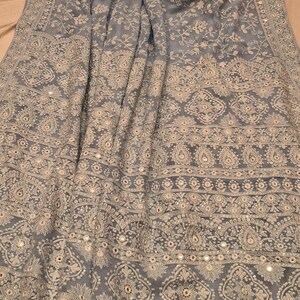 May include: A light blue embroidered shawl with a floral pattern and silver embellishments. The shawl has a border with a repeating design and tassels on the edges.
