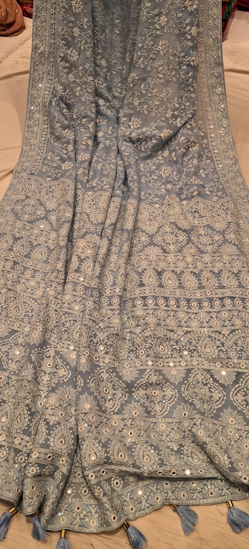 May include: A light blue embroidered shawl with a floral pattern and silver embellishments. The shawl has a border with a repeating design and tassels on the edges.