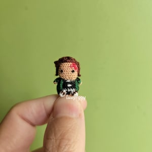 May include: A small, crocheted figure of a character with brown hair, a scar, and a black and green outfit. The figure is held in a hand against a green background. The text "luvpiggy" is visible on the figure.