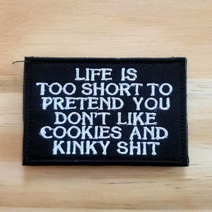 May include: A black rectangular patch with white embroidered text that reads: "LIFE IS TOO SHORT TO PRETEND YOU DON'T LIKE COOKIES AND KINKY SHIT". The patch is on a light brown wooden surface.