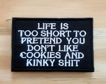 May include: A black rectangular patch with white embroidered text that reads: "LIFE IS TOO SHORT TO PRETEND YOU DON'T LIKE COOKIES AND KINKY SHIT". The patch is on a light brown wooden surface.