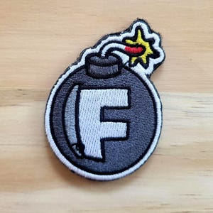 May include: Embroidered patch featuring a stylized bomb design. The patch is primarily black and gray, with a white border and a large white letter "F" in the center. The fuse is red and yellow. The patch is likely for clothing or accessories.