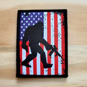 May include: A rectangular patch featuring a silhouette of Bigfoot holding a rifle, set against a distressed American flag design. The flag has red and white stripes with a blue field of white stars. The patch is bordered with black.