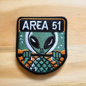 May include: Embroidered patch featuring an alien with large black eyes and a green face. The patch has the text "AREA 51" in white letters on a black background. The alien is set against a green and orange backdrop.