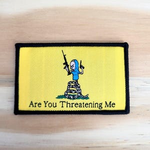 May include: Yellow patch with a black border featuring a cartoon figure holding a rifle and a pistol, atop a coiled snake. The text "Are You Threatening Me" is printed below the image.