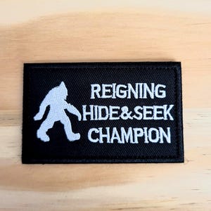 May include: Black rectangular patch with white text and a white silhouette of a Bigfoot. The text reads "REIGNING HIDE&SEEK CHAMPION". The patch is likely for a jacket, backpack, or other apparel.