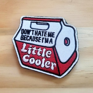 May include: A red, white, and black embroidered patch in the shape of a cooler. The patch features the text "DON'T HATE ME BECAUSE I'M A Little Cooler" in white lettering. The patch has a black border and is set against a wooden background.