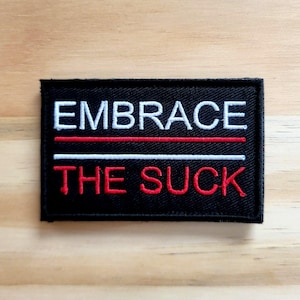 May include: Black rectangular patch with the words "EMBRACE THE SUCK" in white and red lettering. The text is centered and stacked, with a red line separating the words. The patch is on a light brown wooden surface.
