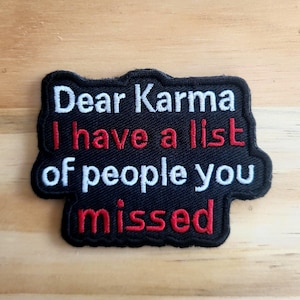 May include: Black embroidered patch with the text "Dear Karma I have a list of people you missed" in white and red lettering. The patch has a black border and is set against a wooden background.
