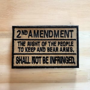 May include: A rectangular patch with the text "2ND AMENDMENT" and the phrase "THE RIGHT OF THE PEOPLE TO KEEP AND BEAR ARMS. SHALL NOT BE INFRINGED." The patch has a tan background with black text and border.