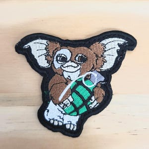 May include: Embroidered patch featuring a brown creature with large ears, holding a green and black grenade. The patch has a black border and white accents on the ears and feet. The creature has a mischievous expression.
