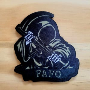 May include: Black patch featuring a Grim Reaper figure holding a rifle. The figure has a skeletal hand and is wearing a hooded cloak. The text "FAFO" is printed in white at the bottom of the patch.