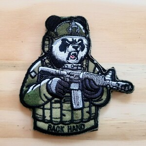 May include: Embroidered patch featuring a panda bear in military gear, holding a rifle. The panda is wearing a helmet and tactical vest. The text "BACK HAND" is visible on the vest. The patch has a green, black, and white color scheme.