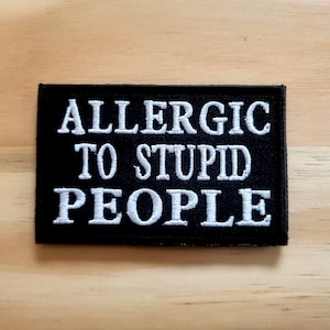 May include: Black rectangular patch with white embroidered text that reads "ALLERGIC TO STUPID PEOPLE". The patch is displayed on a light brown wooden surface.