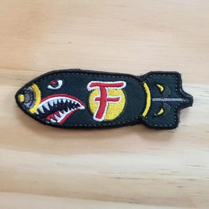 May include: Embroidered patch in the shape of a bomb, featuring a shark mouth design with red and white teeth, a yellow circle with a red "F", and a dark green body. The patch is on a light brown wooden surface.