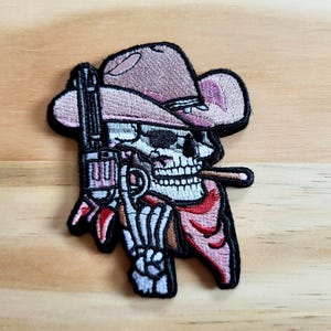May include: Embroidered patch featuring a skull wearing a pink cowboy hat and a red bandana. The skull is holding a revolver. The patch has a black border and is set against a wooden background.