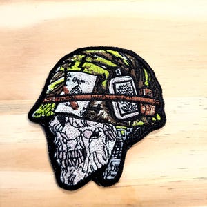 May include: An embroidered patch featuring a skull wearing a camouflage helmet. The helmet is adorned with a playing card, a small rectangular item, and a brown band. The skull is white with black detailing, and the helmet has green and brown tones.