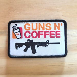May include: A rectangular patch with a black border features the words "GUNS N' COFFEE" in orange and red. A black rifle graphic is below the text, and a coffee cup graphic is in the upper left corner.