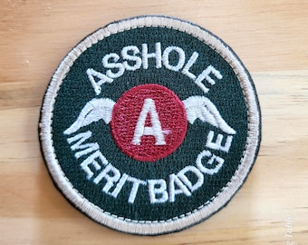 May include: Embroidered patch with the words "ASS HOLE MERIT BADGE" in white lettering on a dark green background. A red circle in the center features a white "A" and white wings. The patch has a beige border.