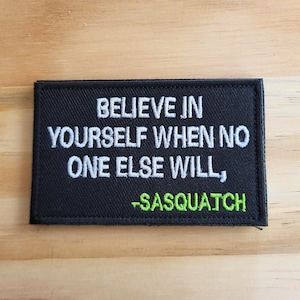 May include: Black rectangular patch with white embroidered text that reads "BELIEVE IN YOURSELF WHEN NO ONE ELSE WILL," with "-SASQUATCH" in green. The patch is on a light brown wooden surface.