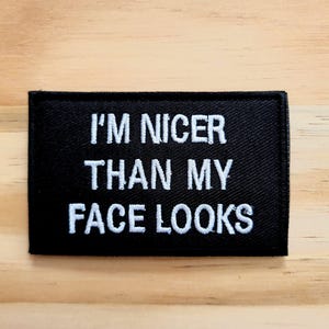 May include: Black rectangular patch with white embroidered text that reads "I'M NICER THAN MY FACE LOOKS". The patch is on a light brown wooden surface.