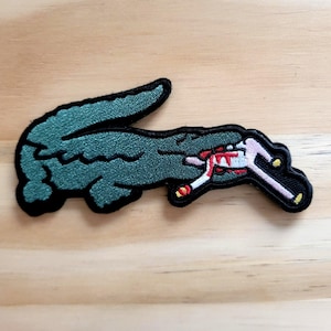 May include: Embroidered patch featuring a green crocodile with a black outline. The crocodile is biting a pink and white object with red accents. The patch is on a light brown wooden surface.