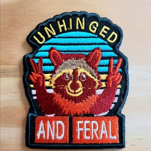 May include: Embroidered patch featuring a raccoon with a peace sign gesture. The patch has the words "UNHINGED" above the raccoon and "AND FERAL" below. The design includes shades of red, brown, yellow, and blue.