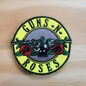 May include: Embroidered patch featuring the Guns N' Roses logo. The design includes a gray center with a gun and floral elements, surrounded by a yellow border with the band's name in black lettering. Two red roses are on either side of the gun.