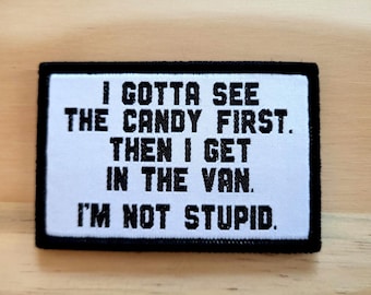 May include: A rectangular patch with black trim and white background. The patch has the text "I GOTTA SEE THE CANDY FIRST. THEN I GET IN THE VAN. I'M NOT STUPID." in black, block letters. The patch is on a light brown wooden surface.