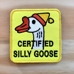 May include: A yellow square patch with a cartoon goose wearing a red and yellow hat. The goose has a red beak and the text "CERTIFIED SILLY GOOSE" in black letters.