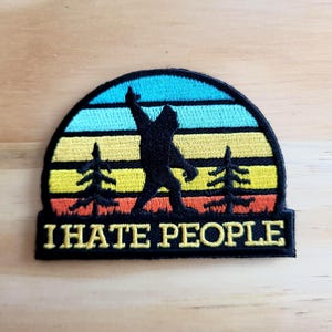 May include: Embroidered patch featuring a black silhouette of Bigfoot with arms raised, set against a sunset design in blue, turquoise, yellow, and orange. The text "I HATE PEOPLE" is displayed below the image.