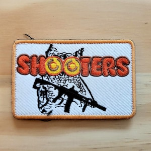 May include: Embroidered patch with the word "SHOOTERS" in orange block letters. The patch features a stylized image of a rifle and a target. The patch has an orange border and is on a white background.