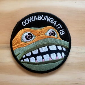 May include: Round embroidered patch featuring a cartoon turtle face with an orange mask and green skin. The patch has the phrase "COWABUNGA IT IS" embroidered in white on a black background. The turtle's mouth is open, revealing white teeth.