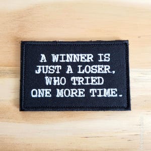 May include: Black rectangular patch with white embroidered text. The text reads: "A WINNER IS JUST A LOSER, WHO TRIED ONE MORE TIME." The patch is on a light brown wooden surface.